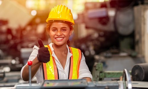Girl teen worker with safety helmet show thumb up working as labor in industry factory with steel machine.